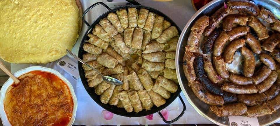 Vedere de sus cu mămăligă, fasole zdrobită, o tigaie mare cu sarmale și o tavă cu cârnați prăjiți, așezate pe o masă tradițională.