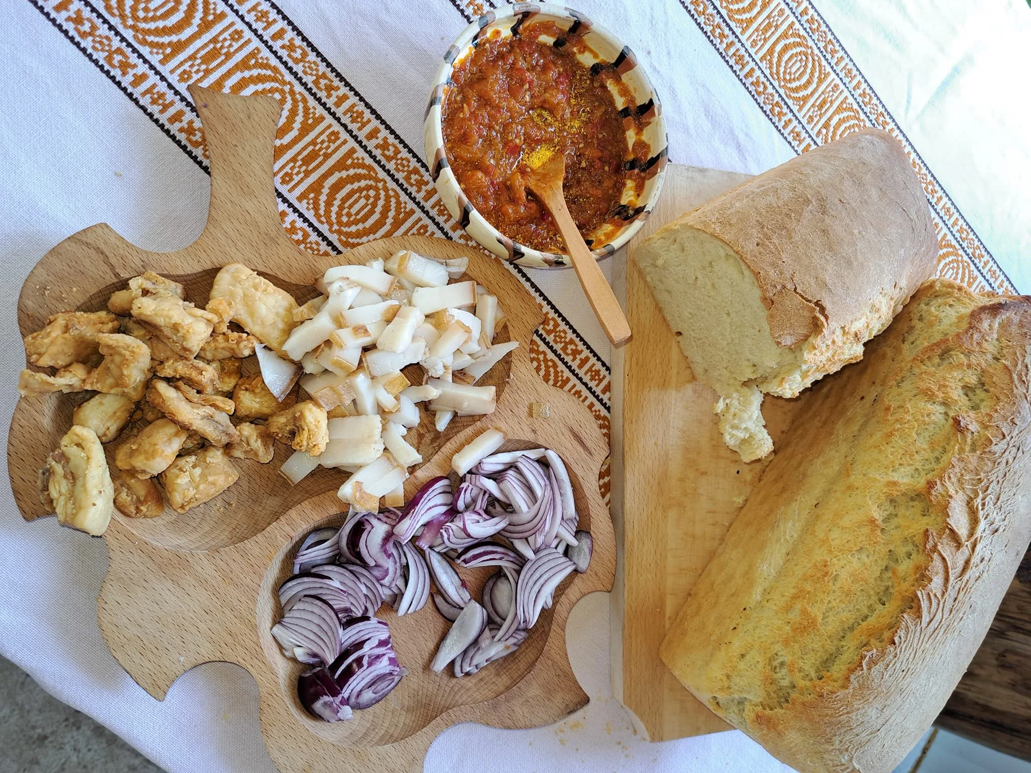 Platou de lemn cu jumări, slănină tăiată cubulețe și ceapă roșie, lângă un castron cu zacuscă și o pâine mare de casă, pe o față de masă tradițională.